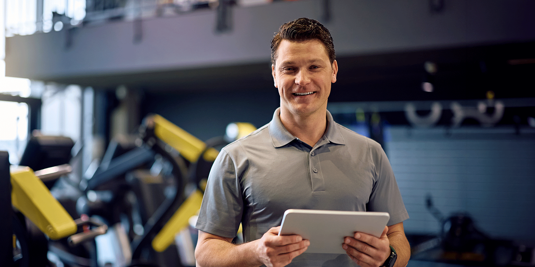 Smiling man in a gray polo shirt holding a tablet while standing in a fitness center. Exercise equipment with yellow accents is visible in the blurred background, and natural light enters from windows on the left.
