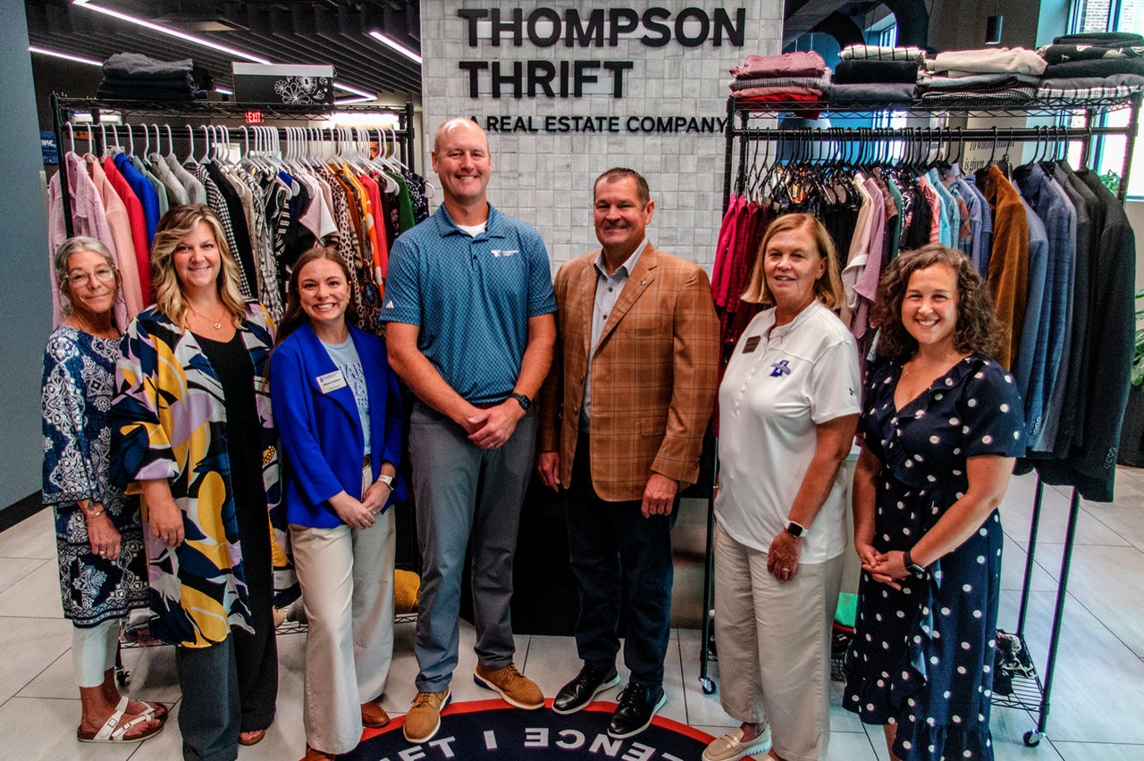 A group of seven people standing in front of clothing racks at Thompson Thrift Real Estate Company. They are gathered on a circular carpet with a visible logo. The background shows various garments on hangers.