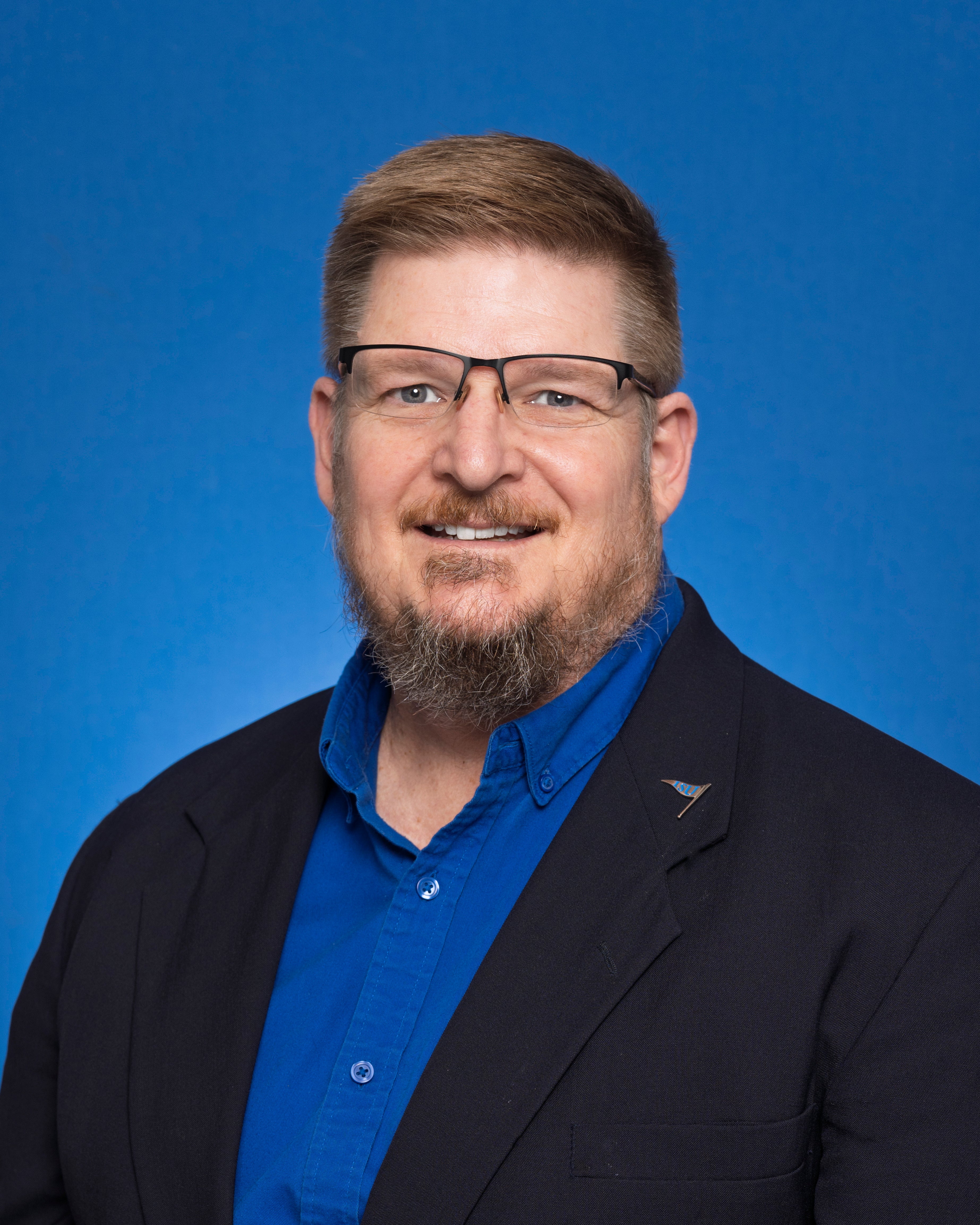 Man with short brown hair, glasses, and a beard wearing a blue shirt and black blazer with an ISU pin on the lapel, against a blue background.