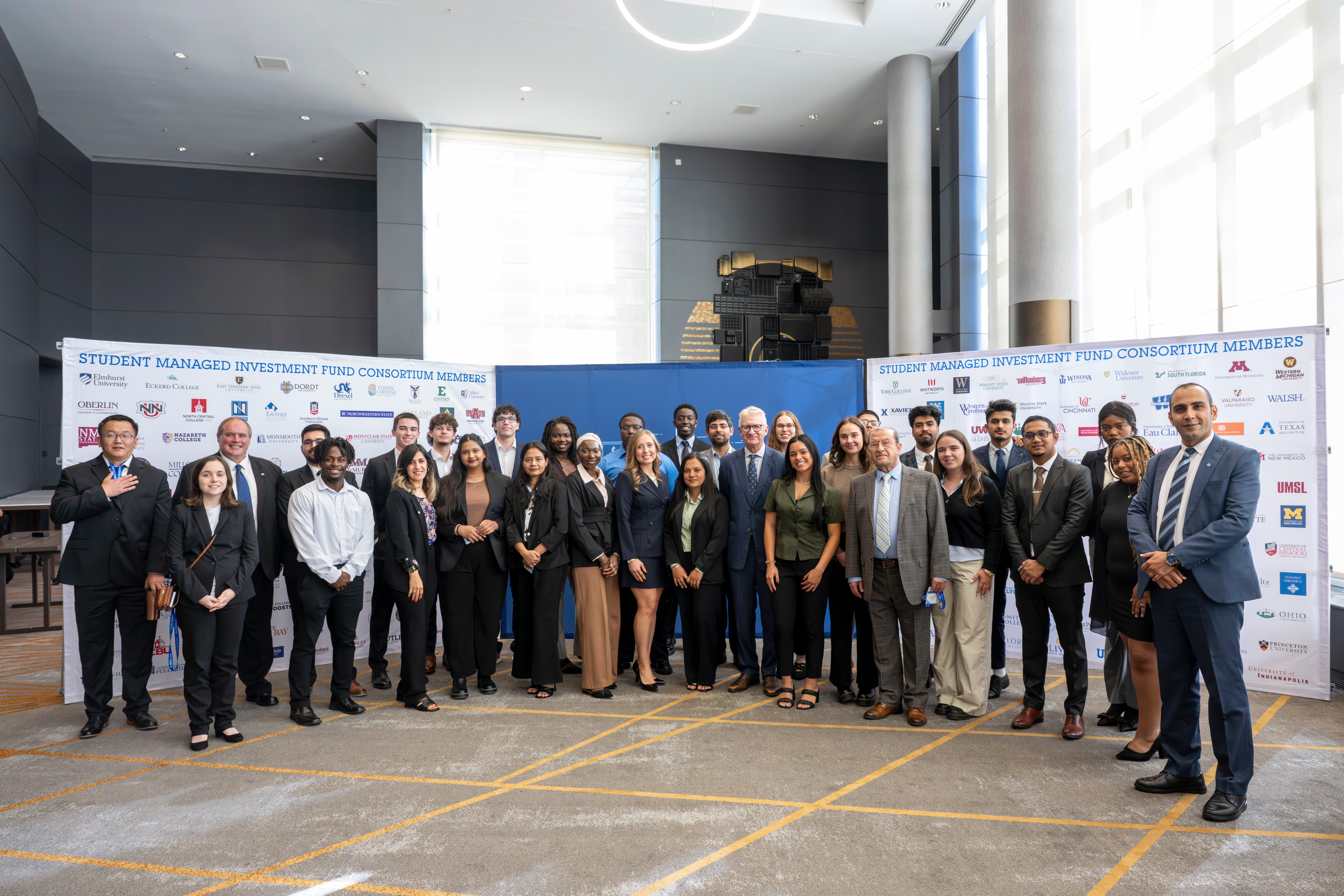 Group of diverse young adults and professionals in business attire standing in front of a conference banner with university logos indoors.