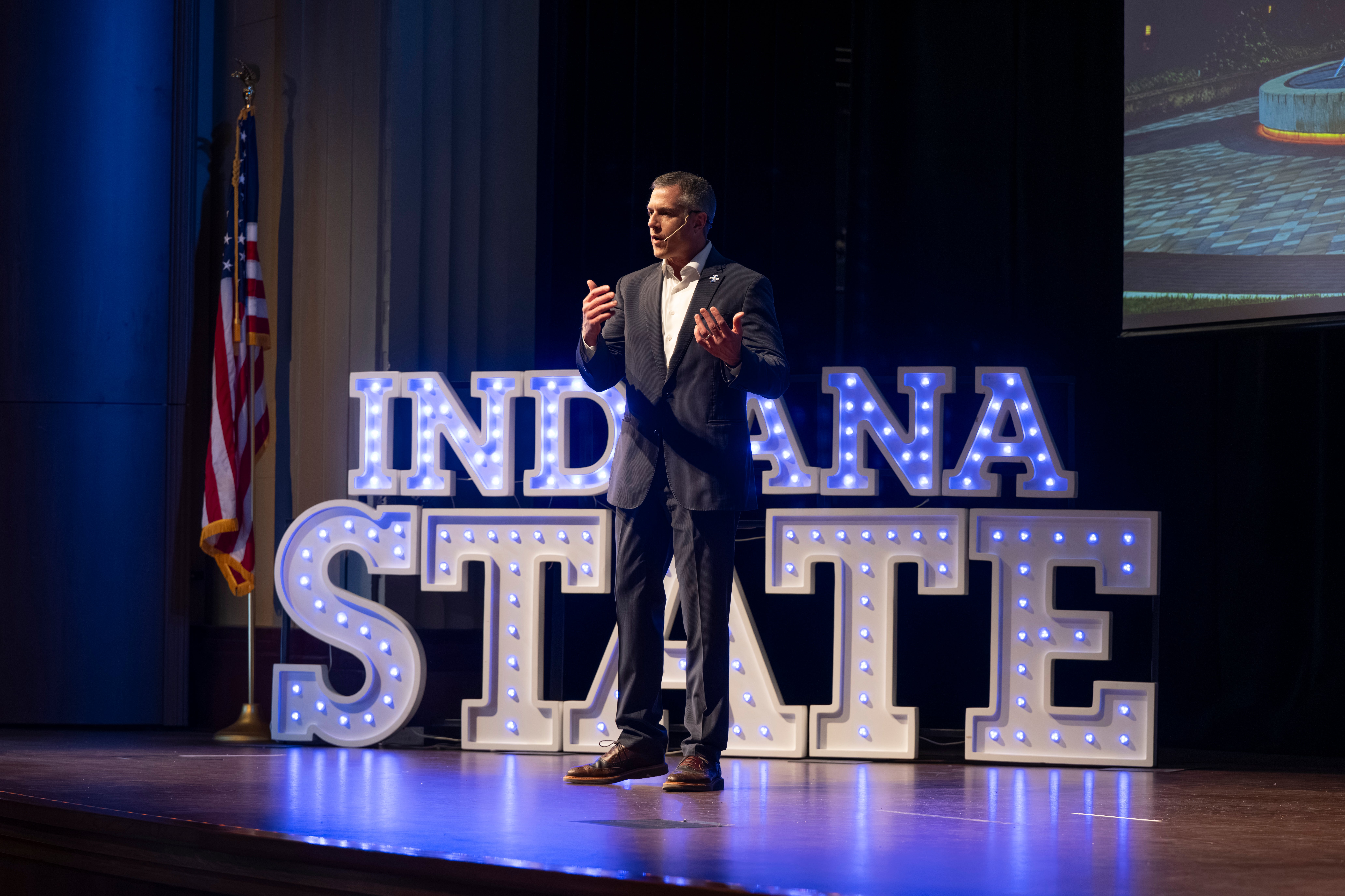 Man in suit speaking on stage with lit Indiana State sign and American flag behind him during university event.