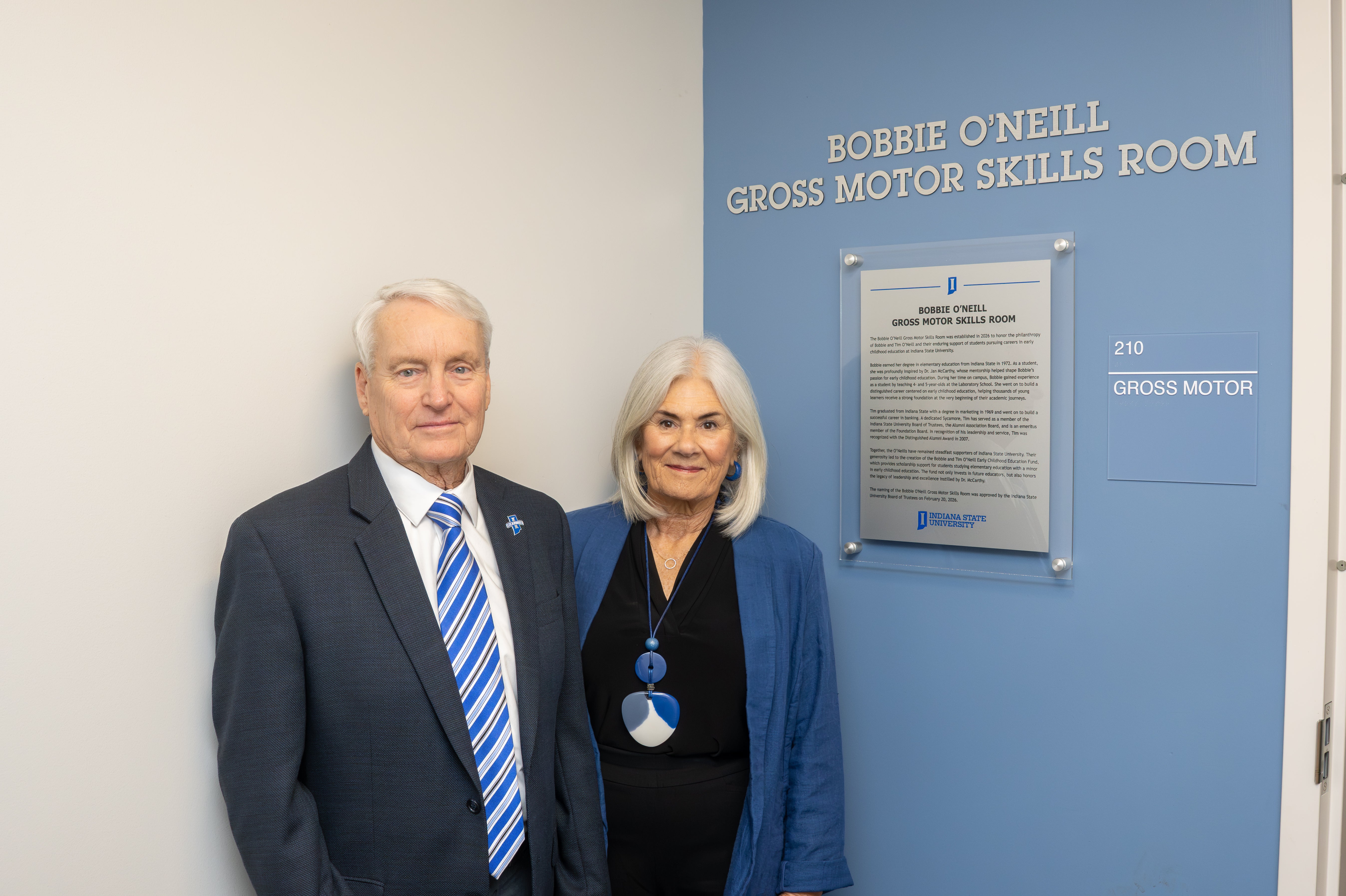 Two people stand beside a blue wall with a sign reading Bobbie O'Neill Gross Motor Skills Room at Indiana State University.