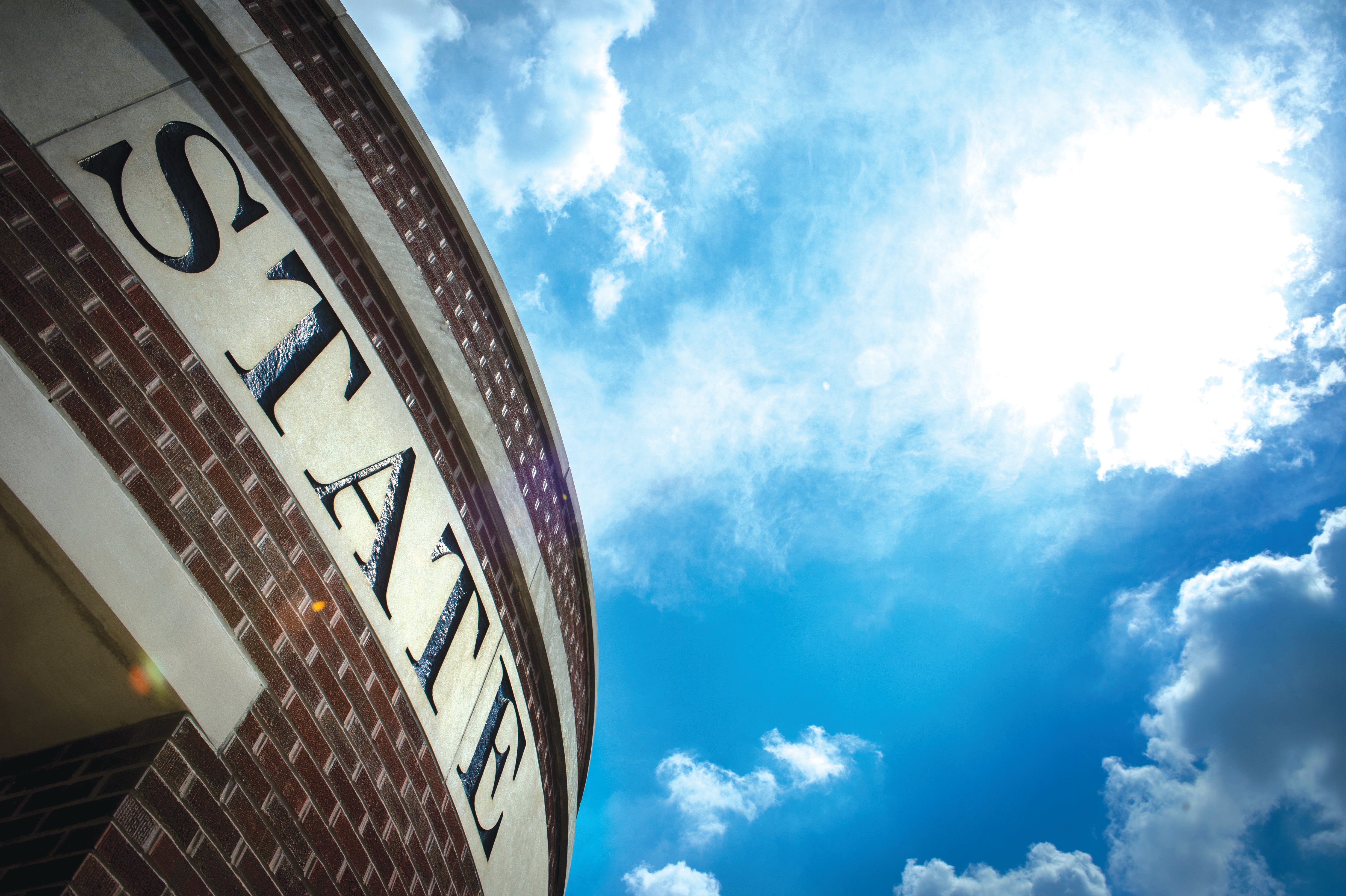 Curved brick building facade with large letters spelling STANE against a bright blue sky with clouds.