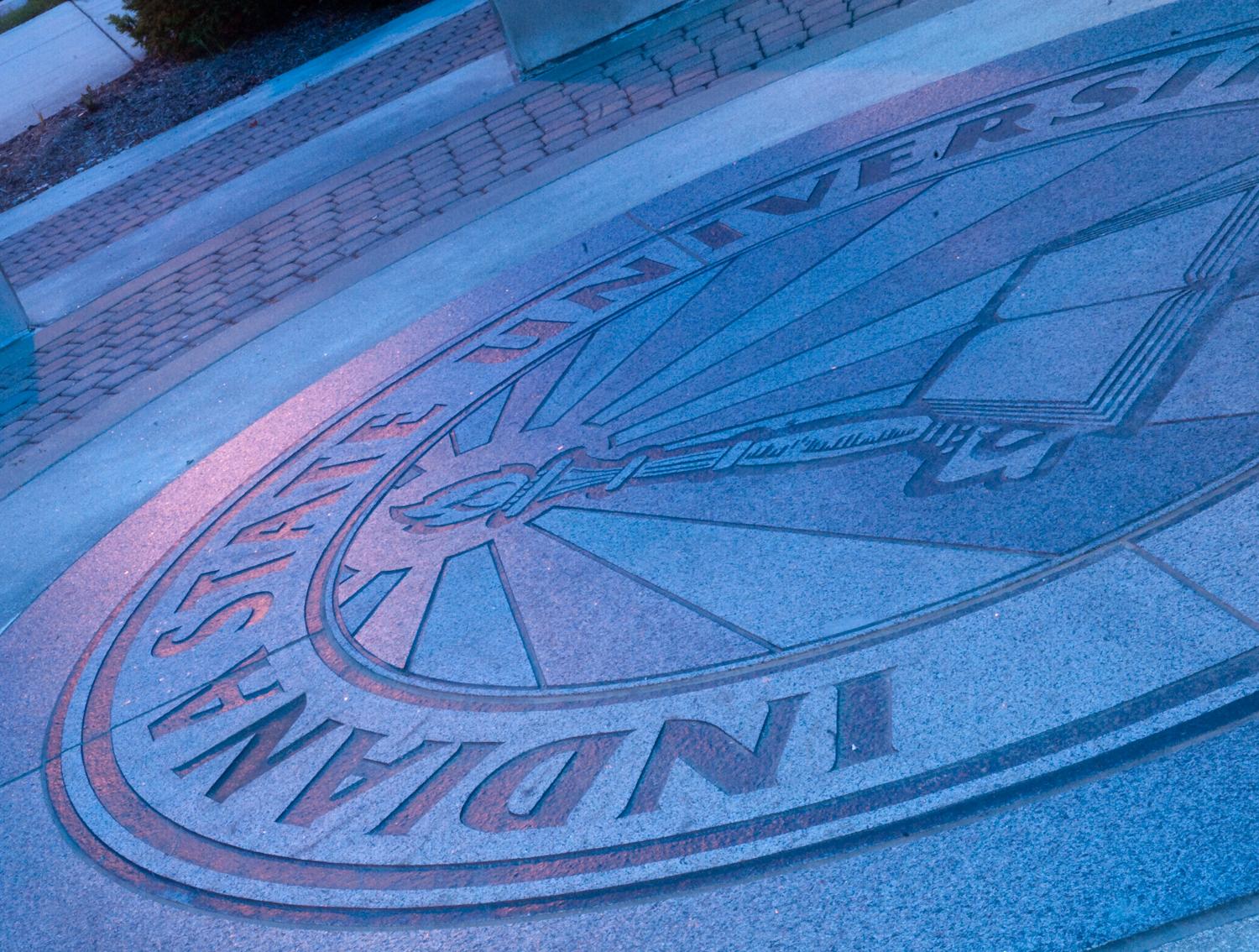 Indiana State University emblem set into outdoor ground with book and torch design surrounded by bricks and pavement.