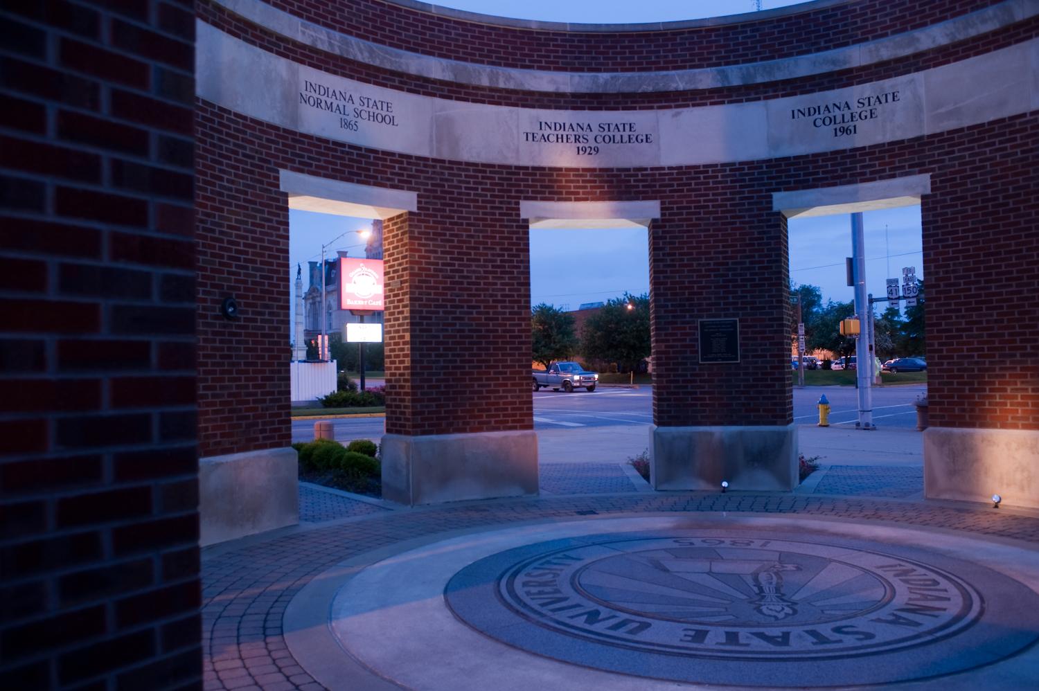 Brick archway with three openings, engraved with Indiana State University historical names and dates, and university seal on the ground at dusk.