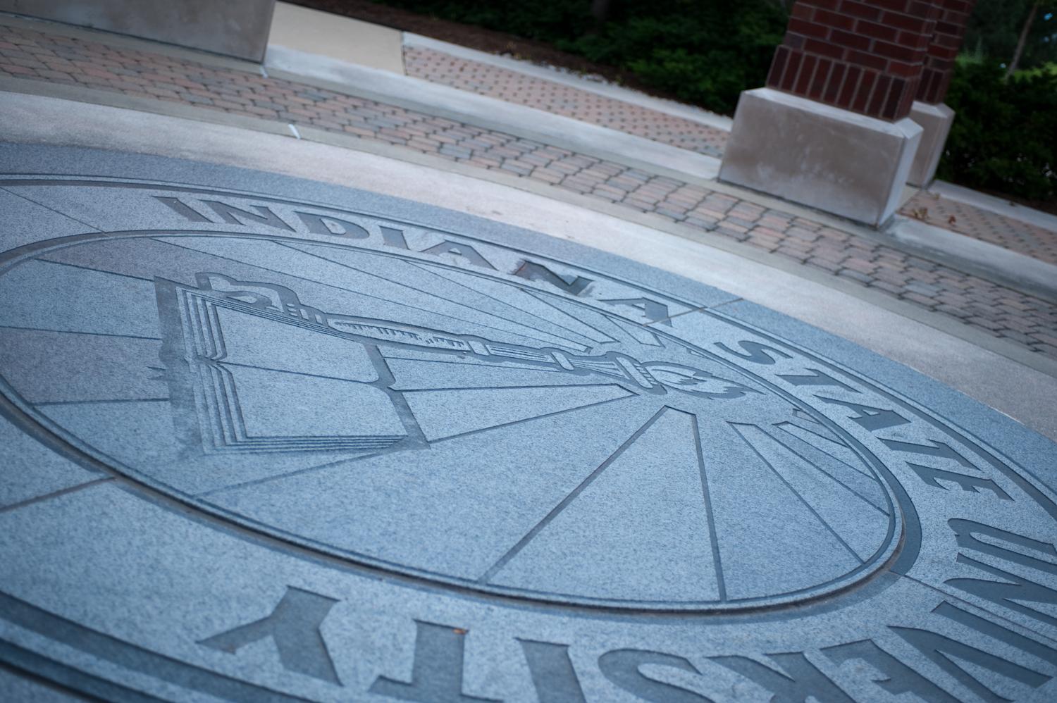 Circular Indiana State University emblem etched in stone ground outside a building entrance with brick columns and greenery.