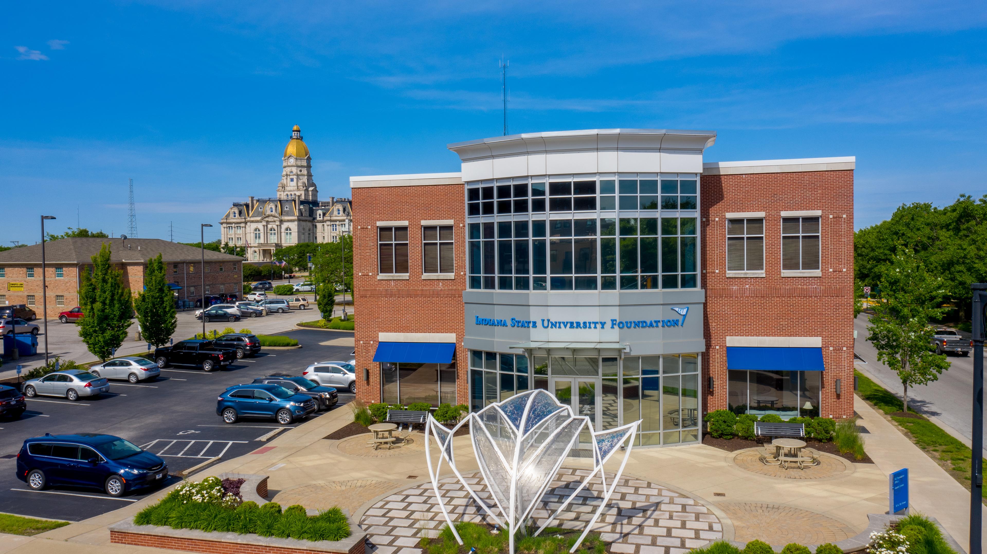 Indiana State University Foundation building with a modern glass entrance, blue awnings, a metal flower sculpture in front, and a historic courthouse with a gol