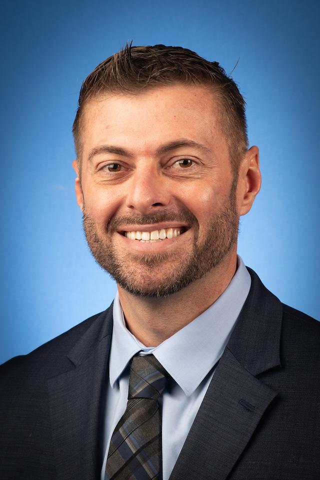 Smiling man with short brown hair and beard wearing a blue shirt, patterned tie, and dark suit jacket against a blue gradient background.