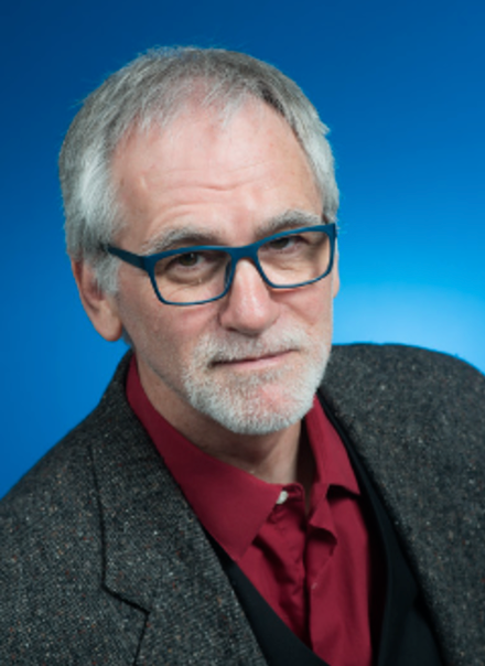 Older man with gray hair, beard, and mustache wearing blue glasses, a red shirt, black vest, and dark blazer against a blue background.