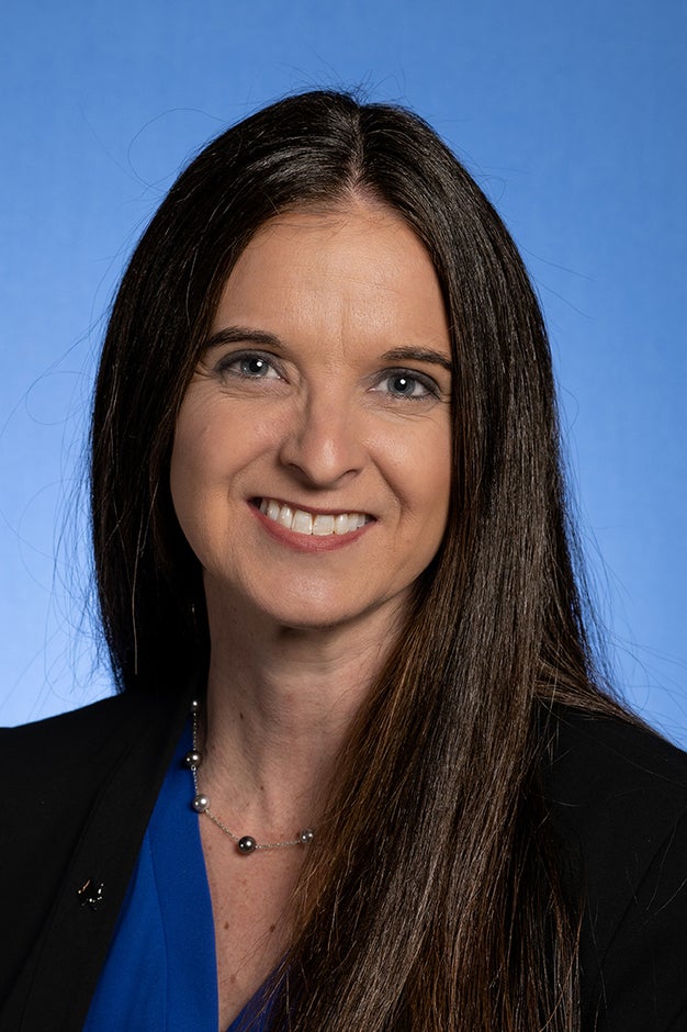 Woman with long straight brown hair wearing a blue top, black blazer, and silver necklace smiling against a blue background.