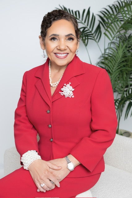 Woman with short curly hair wearing a red blazer adorned with pearl accessories, sitting and smiling against a white background with green plants.