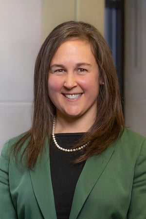 Smiling woman with shoulder-length brown hair, wearing a green blazer, black top, and pearl necklace, standing indoors in front of a wall.