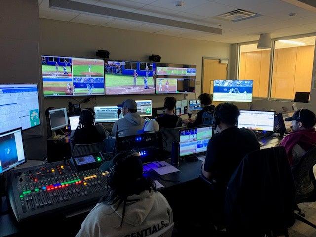 Broadcast production team at work in control room with multiple monitors showing baseball games and audio mixing equipment.