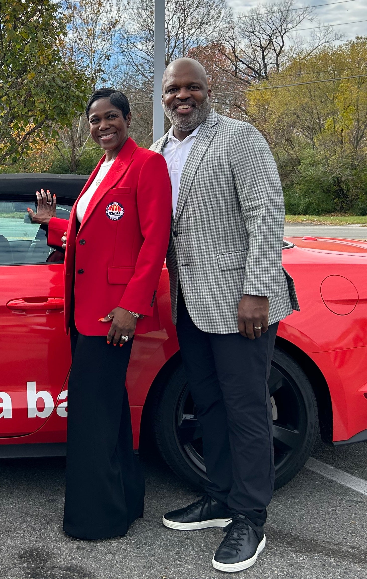 Two people smiling and standing beside a red car, outdoors with trees in the background. One person wears a red blazer with a badge, the other a checked blazer.