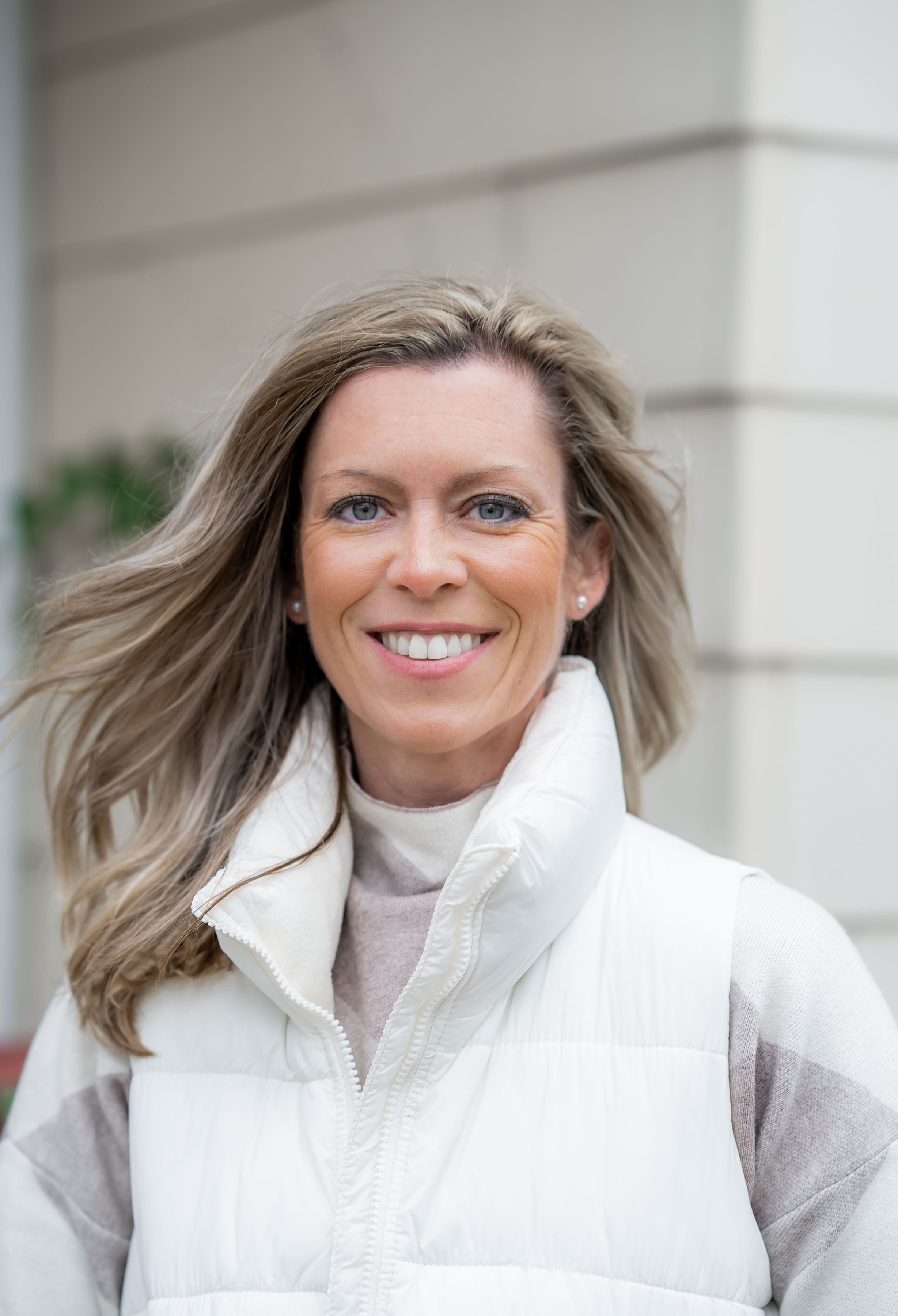 Smiling woman with light brown hair wearing a white puffer vest and striped sweater, standing outdoors near a light-colored building wall.