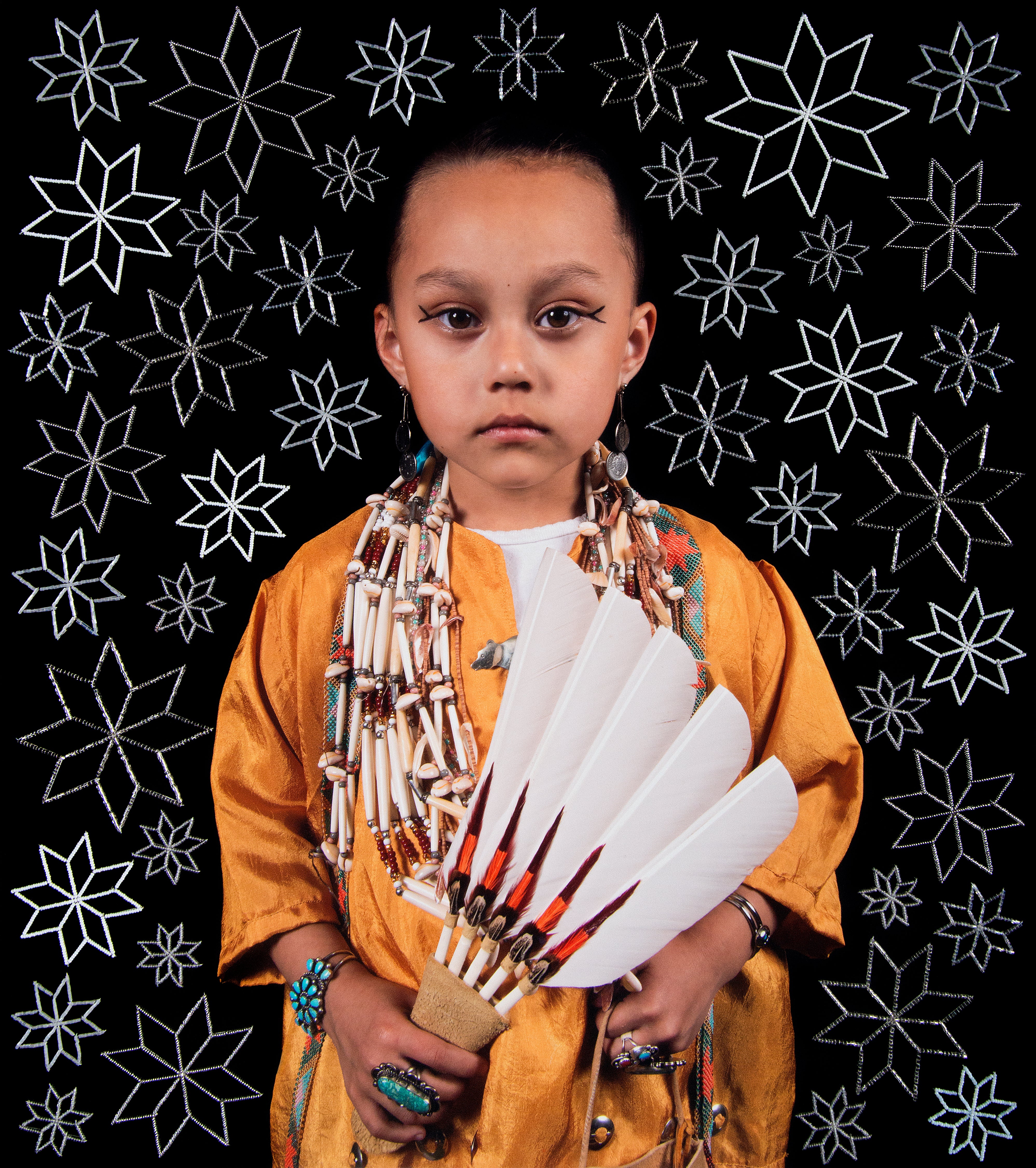 Young Indigenous child in traditional orange garment holds a fan of white feathers with red tips, wearing bead jewelry, against a black background with white st