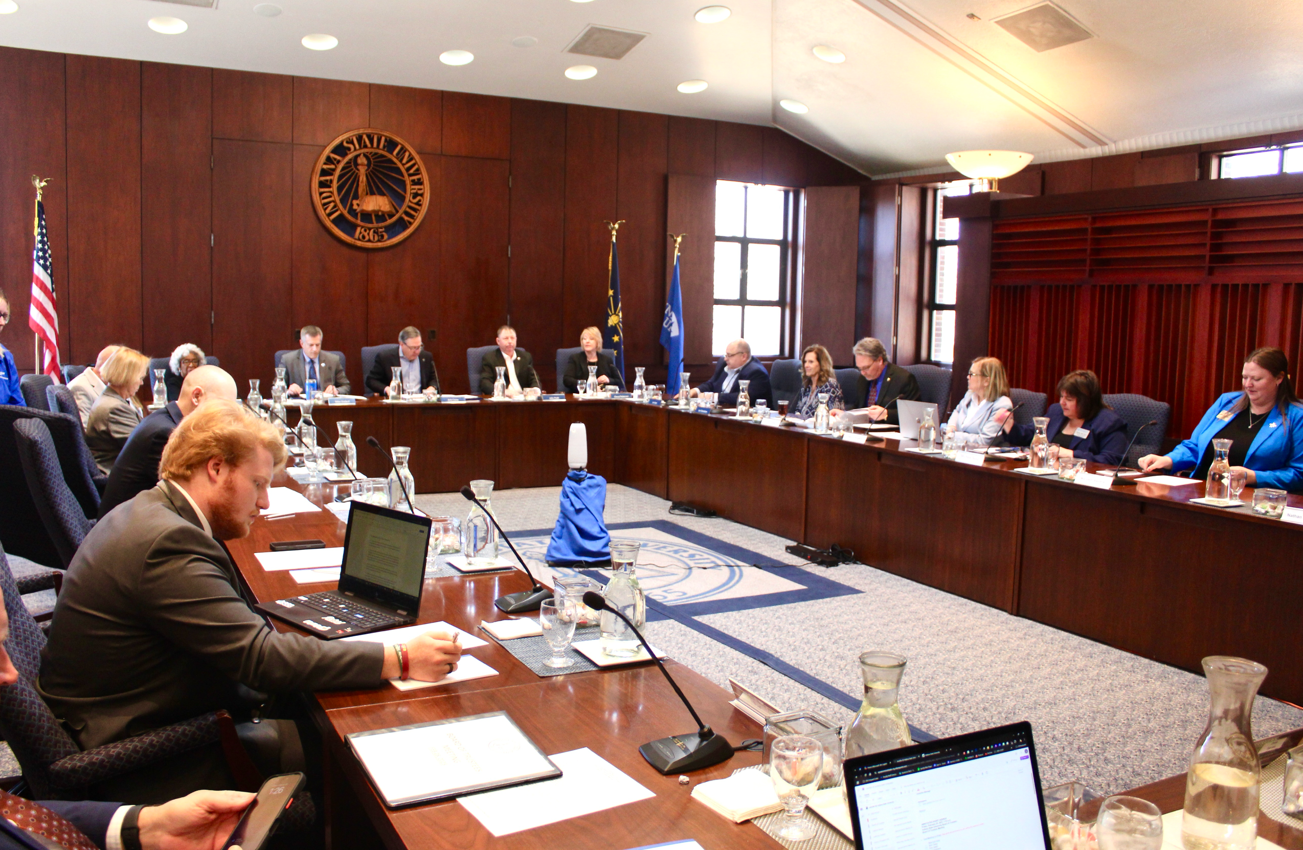 University board members seated around a large U-shaped wood conference table during a meeting in a room with Indiana State University emblem on the wall.
