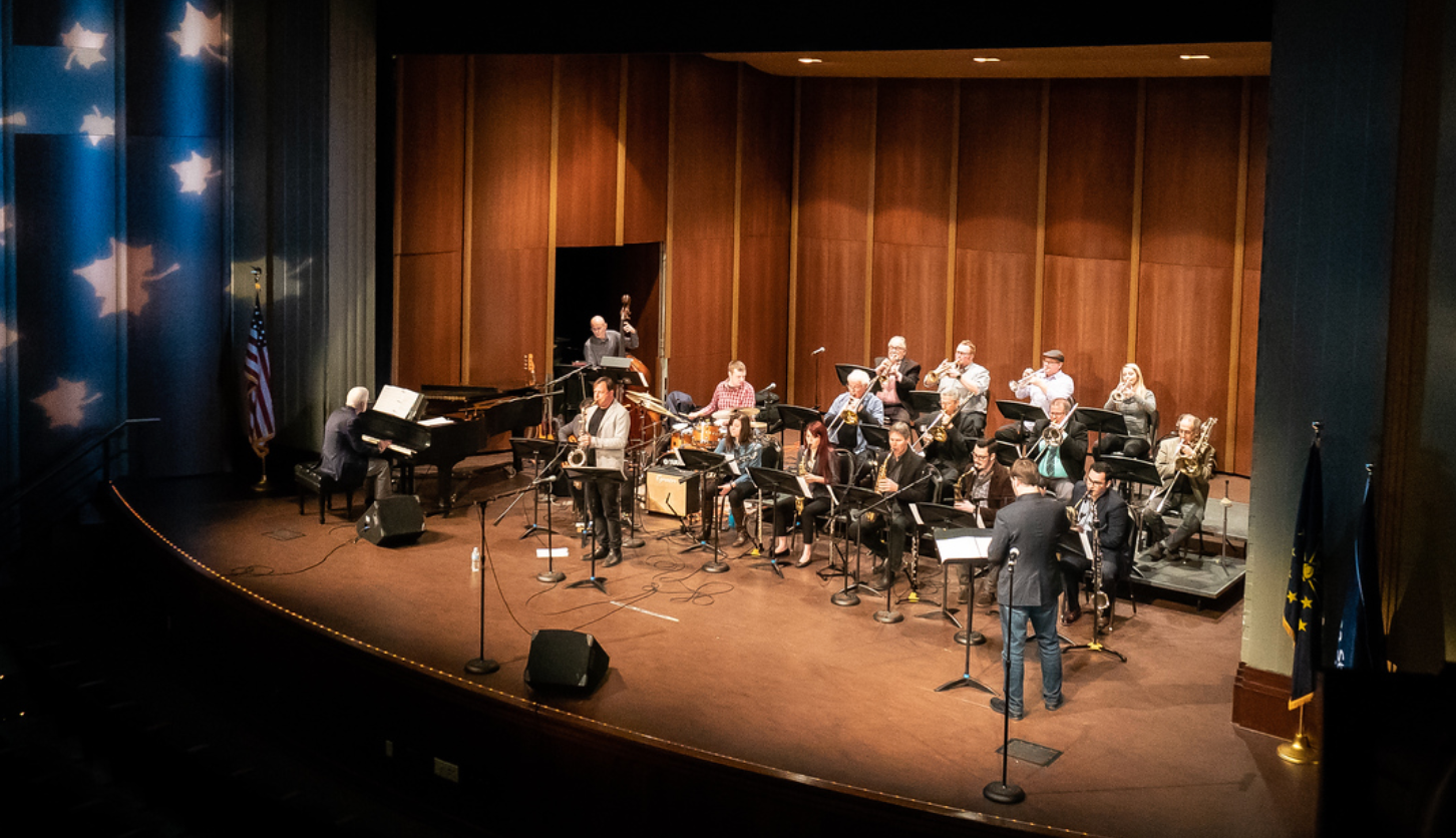 Jazz ensemble performing on stage with piano, saxophones, trumpets, trombones, drums, and a conductor in a large auditorium.