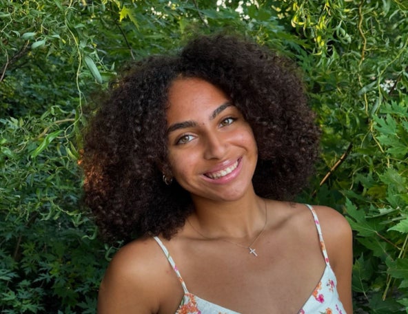 Young woman with curly hair smiling, wearing a floral dress and cross necklace, standing in front of green leafy background outdoors.