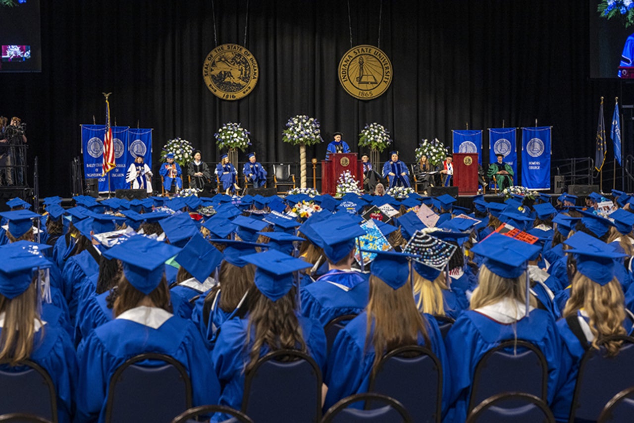 Graduates in blue caps and gowns seated facing a stage with university officials, floral arrangements, and banners at a commencement ceremony.