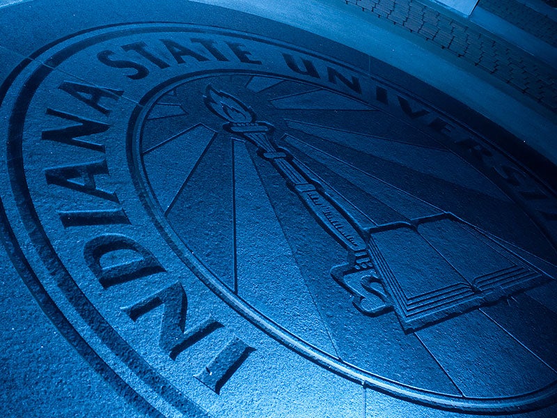 Seal of Indiana State University on pavement, featuring a torch and an open book, with blue lighting.