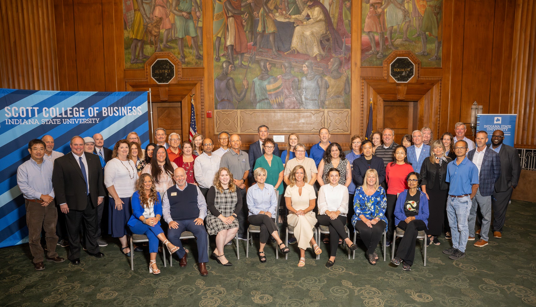 Group of faculty and staff from Indiana State University’s Scott College of Business posing indoors in front of banners and a mural in a large room.