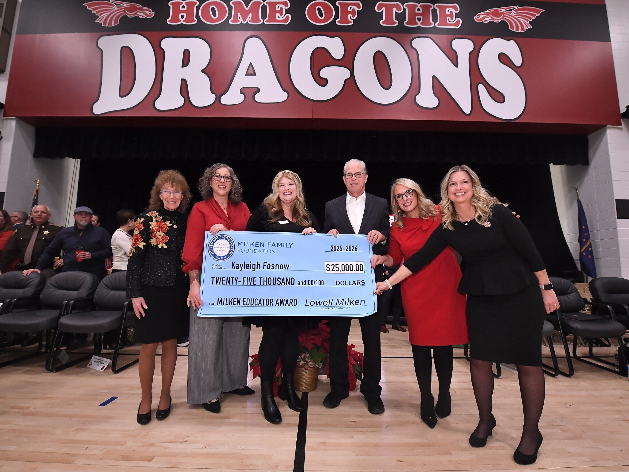 Six people standing in a gym holding an oversized check for $25,000 awarded to Kayleigh Fosnow for the Milken Educator Award.