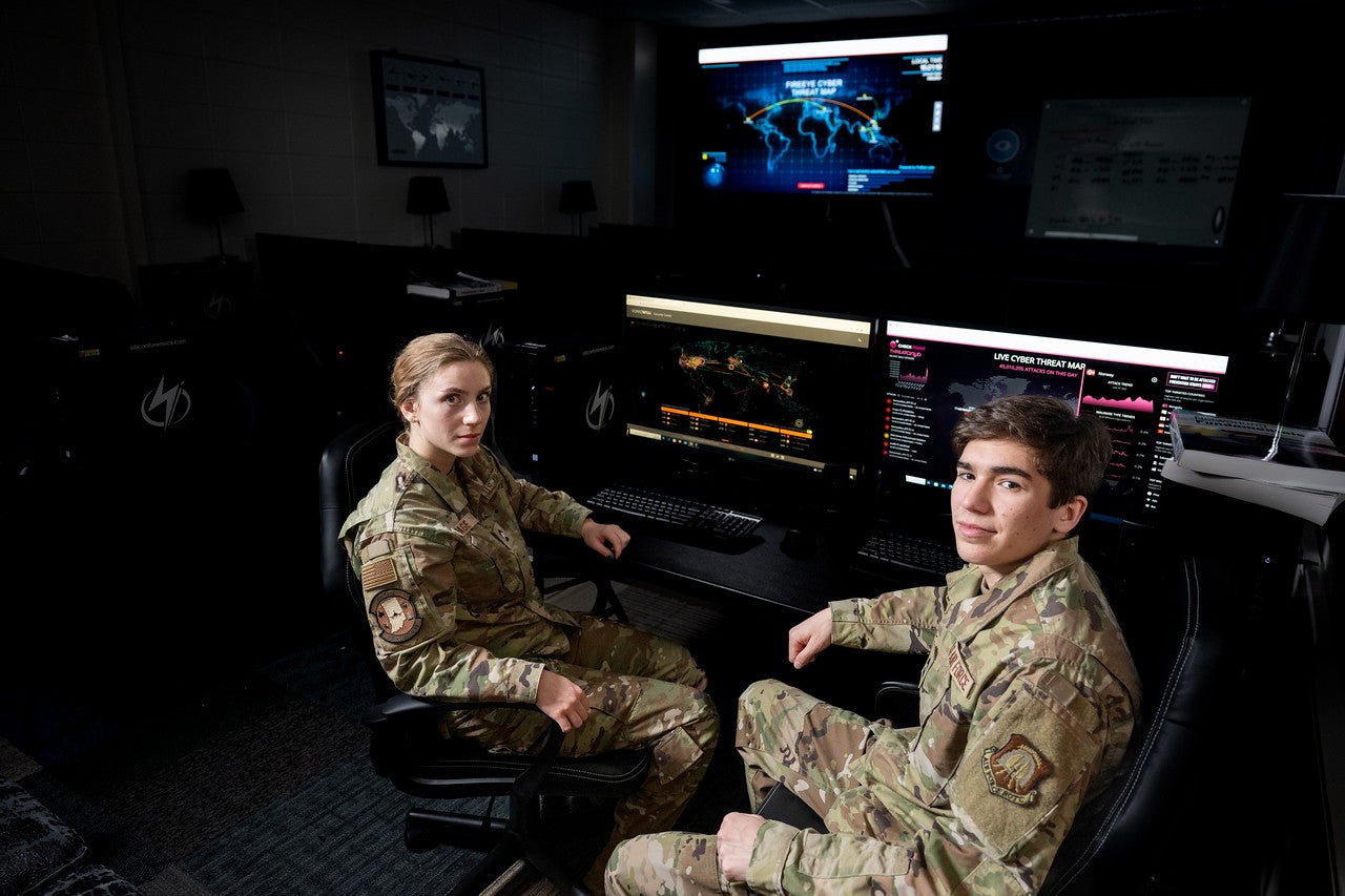 Two military personnel in camouflage uniforms sit in front of computer monitors displaying cyber threat maps in a dimly lit room.