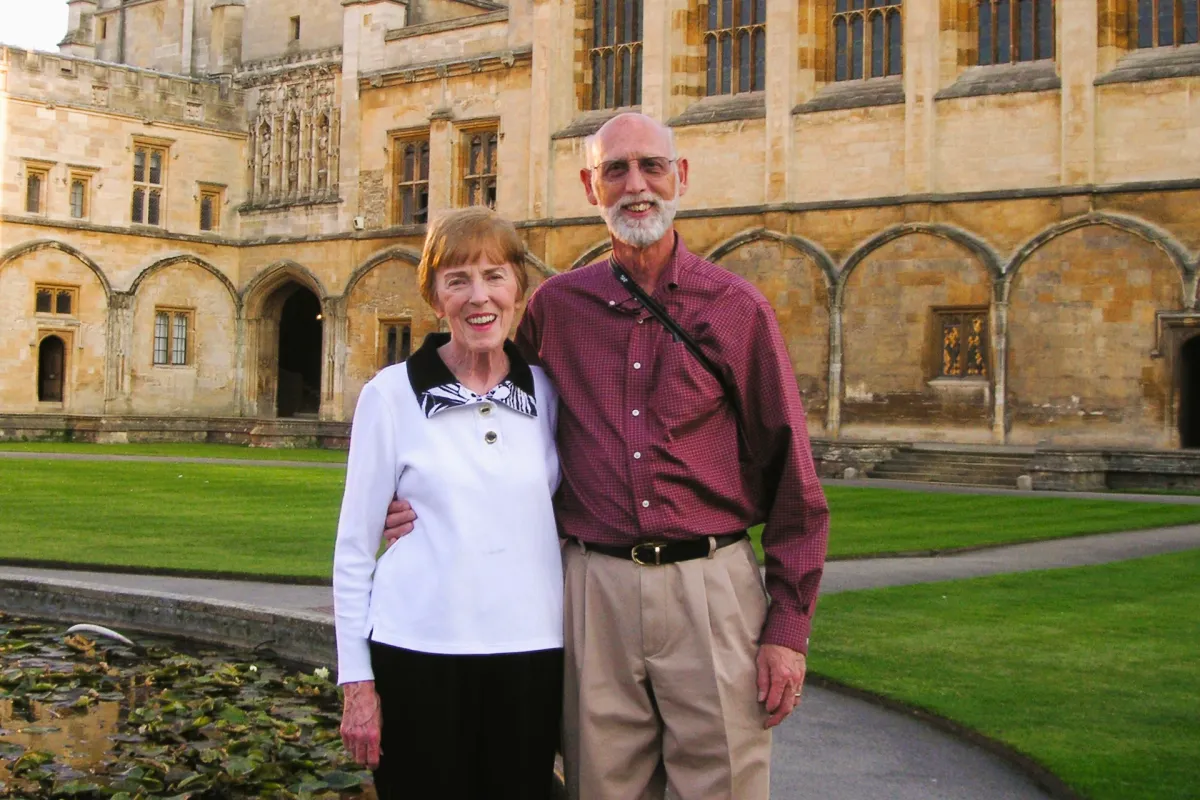 Two people standing together in front of a historic stone university building with gothic architecture and a courtyard pond.