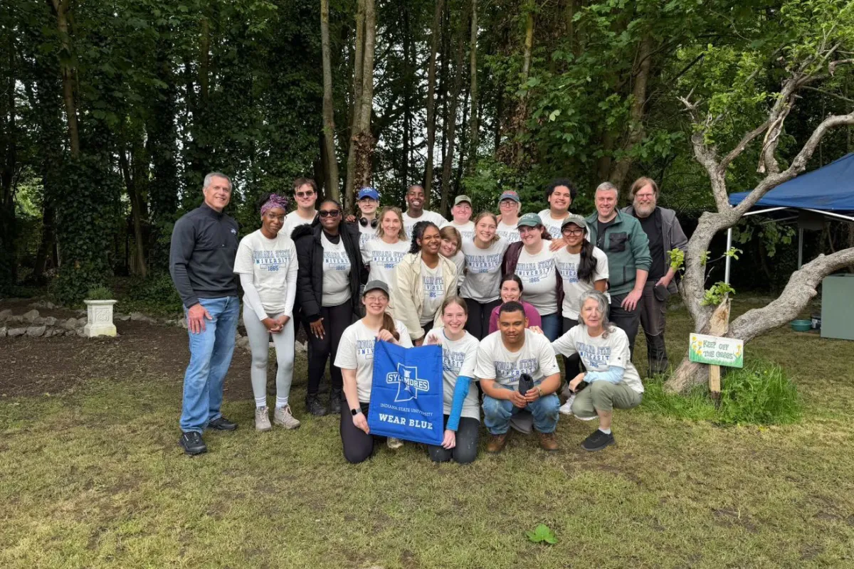 A group of people, some wearing "Wear Blue" shirts, stand and kneel on grass in a wooded area. One person holds a sign with the "Indiana State University: Wear Blue" logo. There's a small tent and a "Plant Sale" sign nearby.