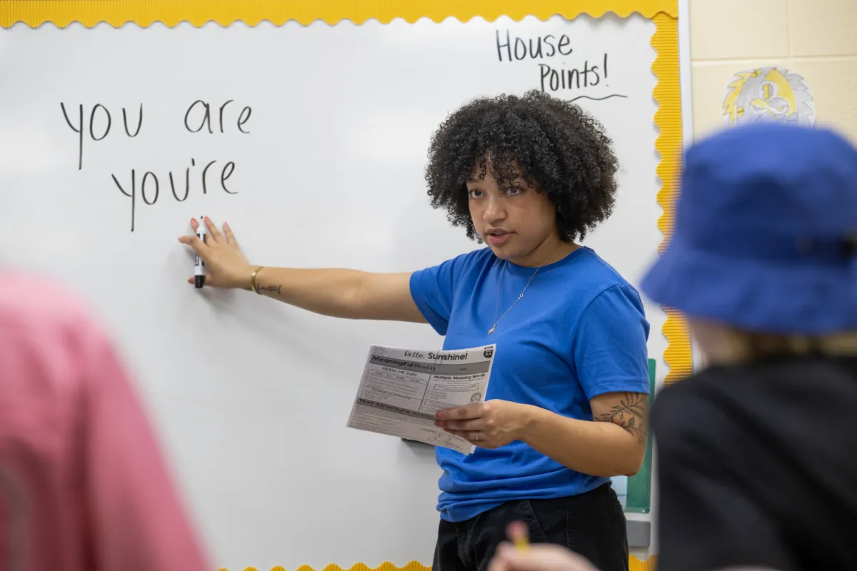 A student teacher stands at a whiteboard, pointing to the words "you are" and "you're" written on it. They hold a paper titled "Worksheet" and are wearing a blue shirt. The board is bordered with yellow paper, and "House Points!" is written in the corner. Two younger children are partially visible in the foreground.