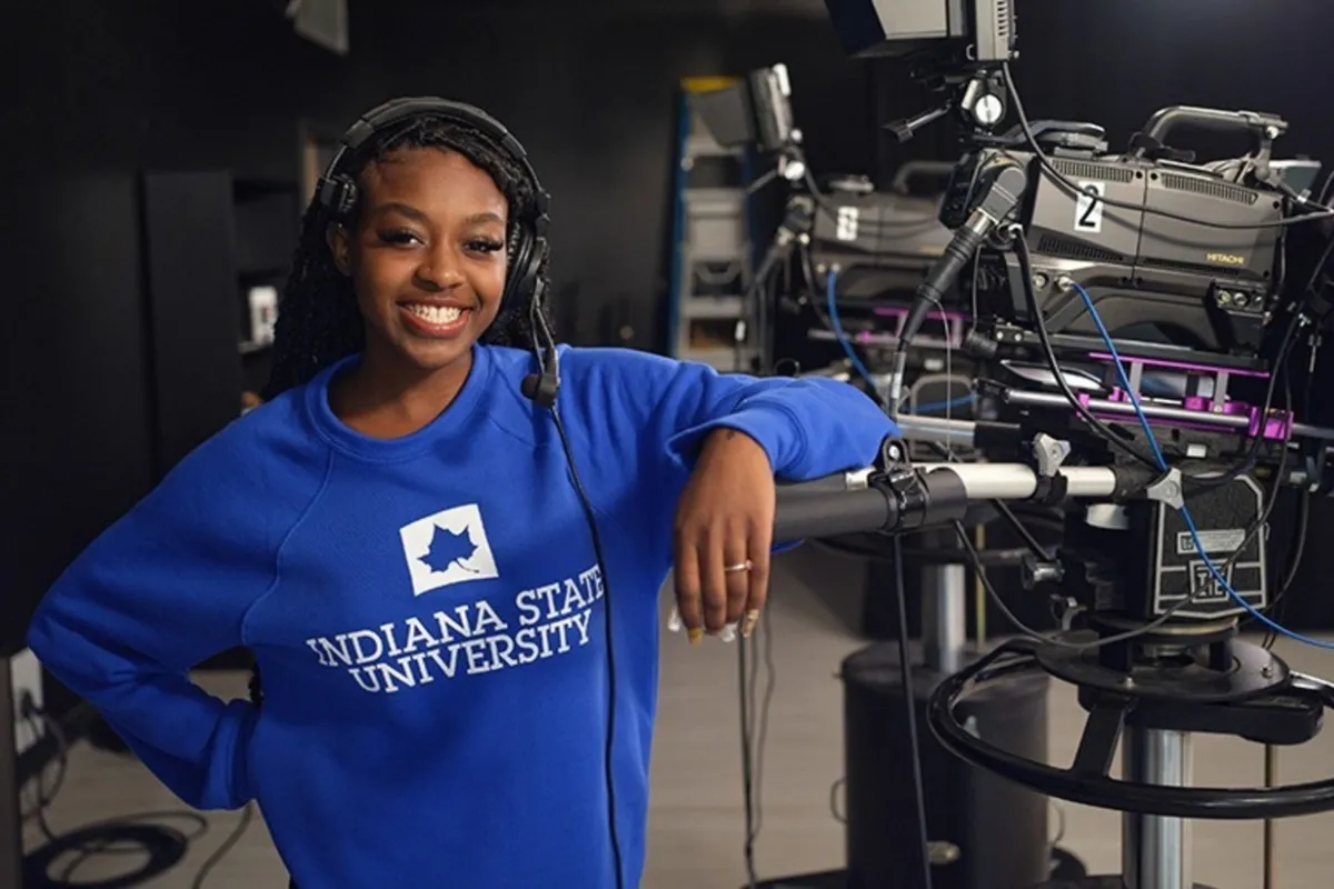 A person in a blue "Indiana State University" sweatshirt, wearing a headset, stands smiling in a television studio next to a large video camera.