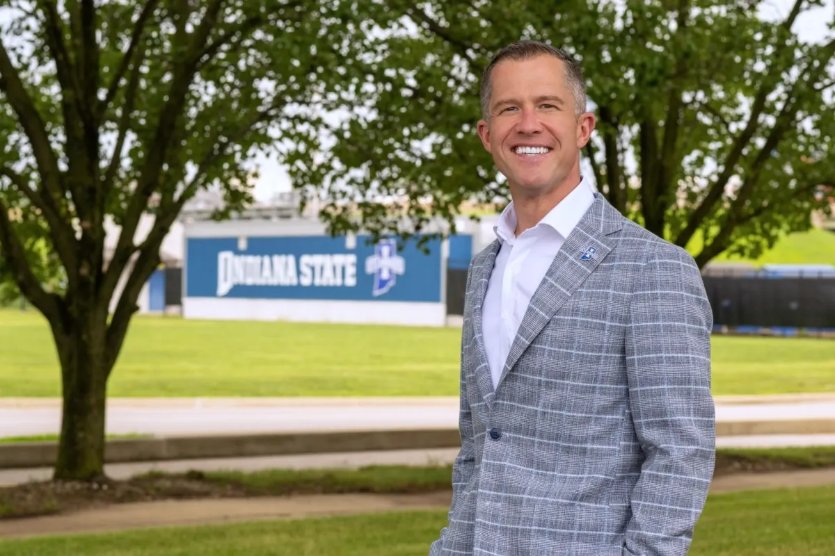 A person in a gray plaid suit stands smiling outdoors with trees and grass in the background. A building with signage that reads "Indiana State" is visible in the distance.
