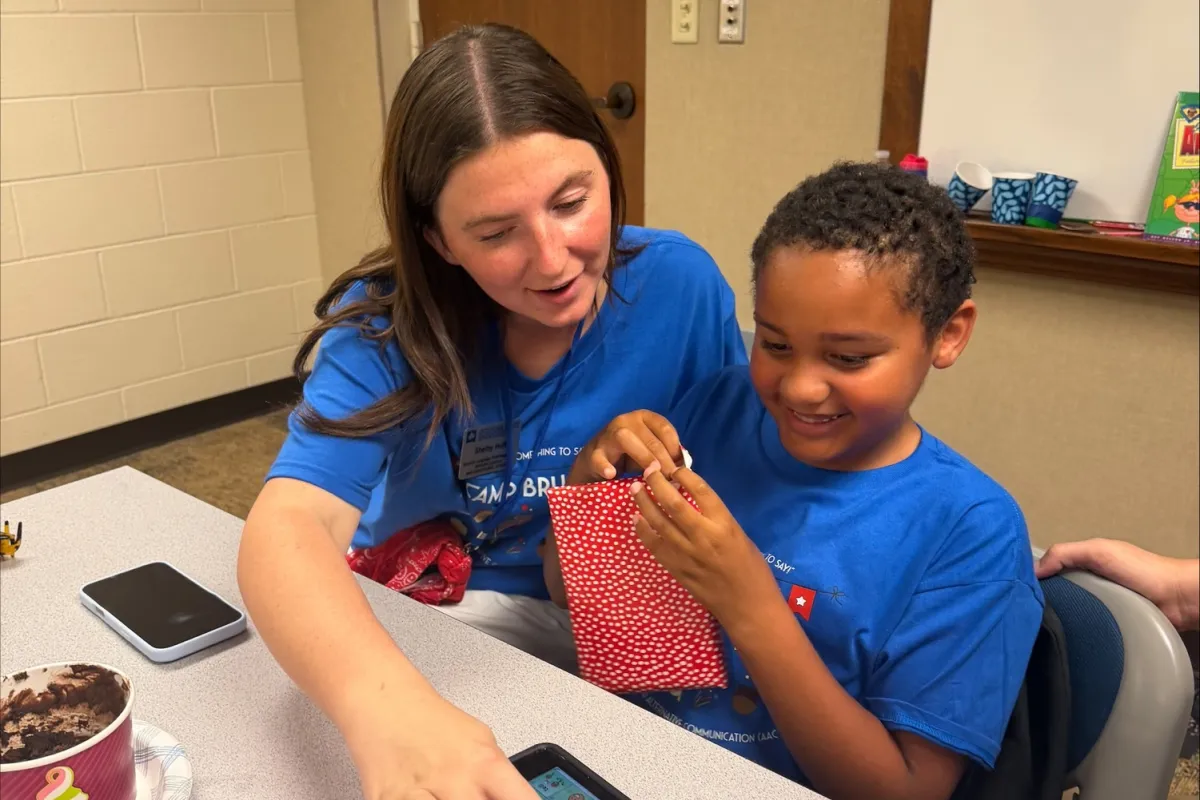 A student assists a young child with an augmentative and alternative communication (AAC) device.