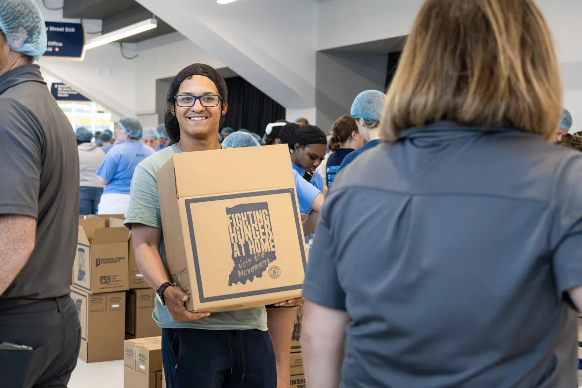 A person carrying a box labeled "Fighting Hunger at Home" at a community event. People in the background are wearing hair nets and sorting boxes.