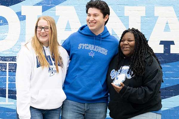 Three students standing in front of a blue-and-white painted Indiana State mural. On the far left is a white female student with straight blonde hair. She wears a white Indiana State sweatshirt, black glasses, and blue jeans. In the middle is a white male student with black hair. He wears a blue Sycamores sweatshirt and blue jeans. On the right is a Black female student with black dreadlocks. She wears a black Sycamores sweatshirt and blue jeans.  