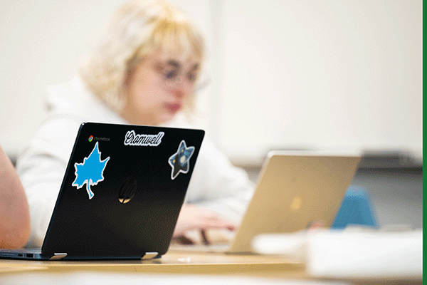 A black laptop is visible in the foreground on a table in a classroom. It has a blue Sycamore leaf sticker with a white outline on the top left, and another stick top center that says Cromwell. A third sticker, further right, is shaped like a star. Out of focus in the background, a blonde female student wearing glasses and a white hoodie can be seen typing on a silver laptop.  