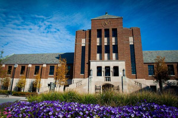 A front view of a large, impressive brick building, known as Tirey Hall. The structure features a central tower with tall vertical windows and decorative stone accents. The entrance is elevated, accessed by a wide staircase flanked by stone railings. The facade includes a circular emblem near the top of the tower and the words "STUDENT UNION BUILDING" inscribed above the entrance. The landscaping in front includes neatly trimmed grass, colorful purple flowers, and small trees with autumn foliage.