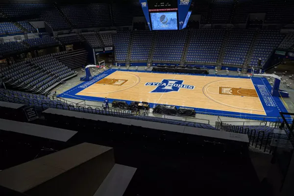 An empty basketball arena viewed from an elevated position, showing a central hardwood court with "Sycamores" and a large blue "I" logo in the middle. The court is surrounded by blue and white markings, with the words "Indiana State University" visible. The seating area around the court consists of tiered rows of blue seats, and a large scoreboard hangs from the ceiling, displaying a Pepsi logo and other information. The arena is well-lit, and there are no spectators present. A few staff members can be seen