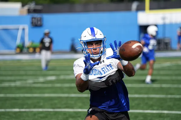 A football player wearing a white helmet with a blue stripe, a white jersey with "Indiana State" written in blue letters, and blue gloves is focused on catching a football. The player is on a green football field with a blurred background of other players and equipment. The player's hands are extended, and the football is just inches away from them, showing intense concentration and athleticism.