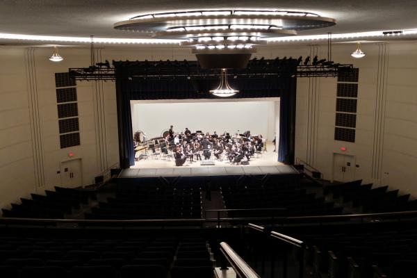 A band performing on stage in Tilson Auditorium, viewed from a distance. The stage is well-lit with musicians seated and playing their instruments, while the auditorium is mostly empty.