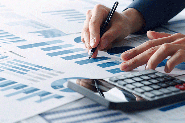 Detail image of a spreadsheet on a table along with a calculator and a person’s hand making notes on the spreadsheet.