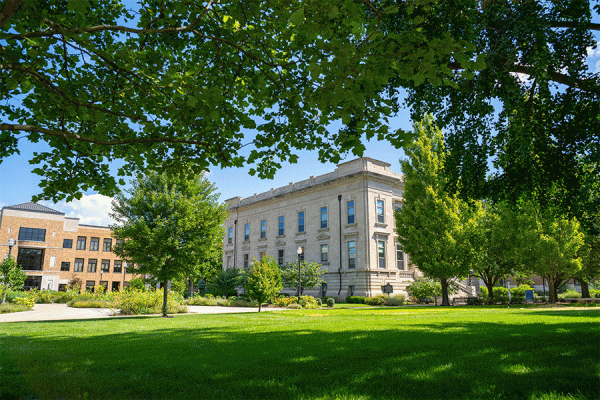 A grey stone building, Normal Hall, is seen at a distance from beneath a canopy of green trees. It is summer, the sun is out, and the grass is green. The reddish-brown brick Science Building is also visible to the left of Normal Hall.