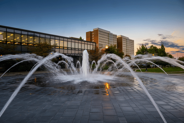 A large fountain with multiple streams of water arching toward the center, situated in front of a modern building labeled “John H. Moore Welcome Center.” The building has many windows. In the background, tall buildings and trees are visible under a clear sky with sunset hues.