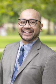 A smiling man wearing round black glasses, a gray suit jacket, a blue striped dress shirt, and a blue patterned tie, standing outdoors with a blurred green park background.
