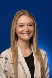 A smiling woman with long straight blonde hair, wearing a light plaid blazer over a black top and a cross necklace, posed against a solid blue background.