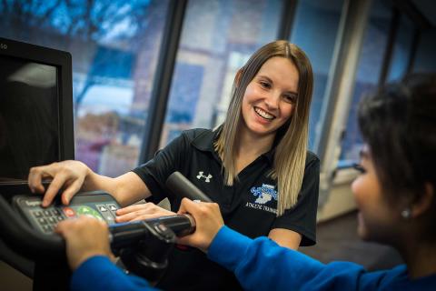 A SMILING FEMALE STUDENT IN A BLUE SWEATER HOLDS ONTO THE HANDLEBARS OF A STATIONARY BIKE WHILE ANOTHER SMILING, FEMALE STUDENT IN A BLACK SYCAMORES ATHLETIC TRAINING POLO SHIRT PUSHES BUTTONS ON THE KEYPAD. A LARGE WINDOW IS VISIBLE BEHIND THEM.
