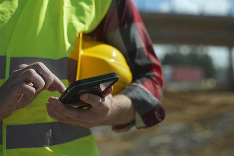 A worker in a yellow safety vest holds a hard hat under their arm while typing on a cell phone