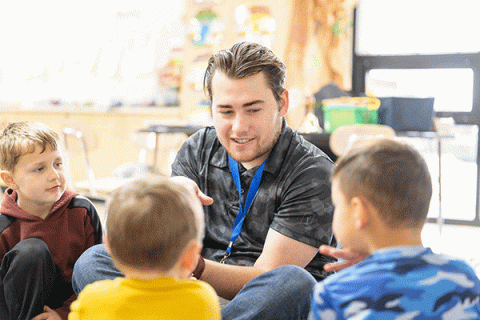 A white, male Indiana State student-teacher, with brown hair and wearing a gray camo t-shirt and blue lanyard, sits and talks with two young boys in a classroom. He is smiling.