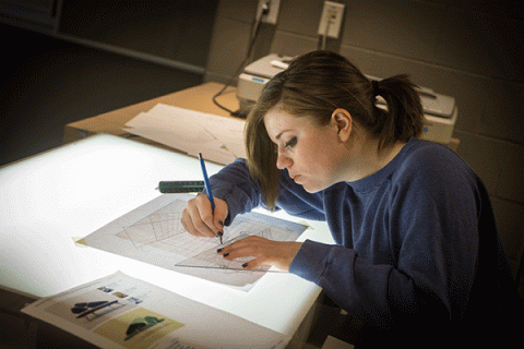 In a lab space, a white female student with brown hair pulled back into a ponytail, sits leaned over an illuminated drafting table. She is wearing a blue long-sleeved shirt. She holds a pencil in her right hand, a protractor in her left hand, and she is drawing a design on a page.